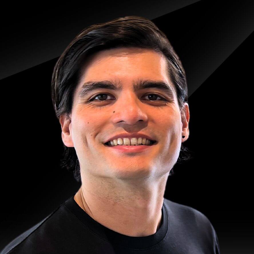 A man with dark hair and a black shirt smiles against a black background.